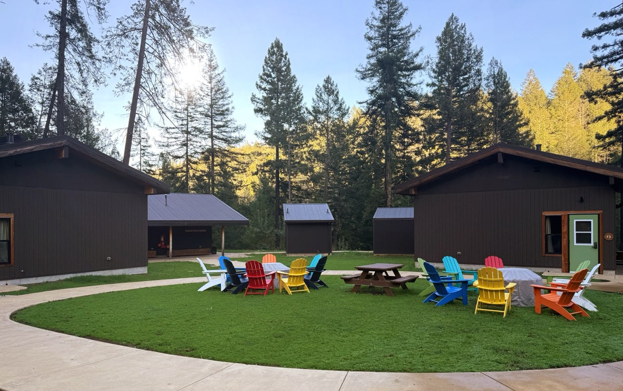 Colorful chairs arranged around a picnic table in a campground with cabins and trees in the background.