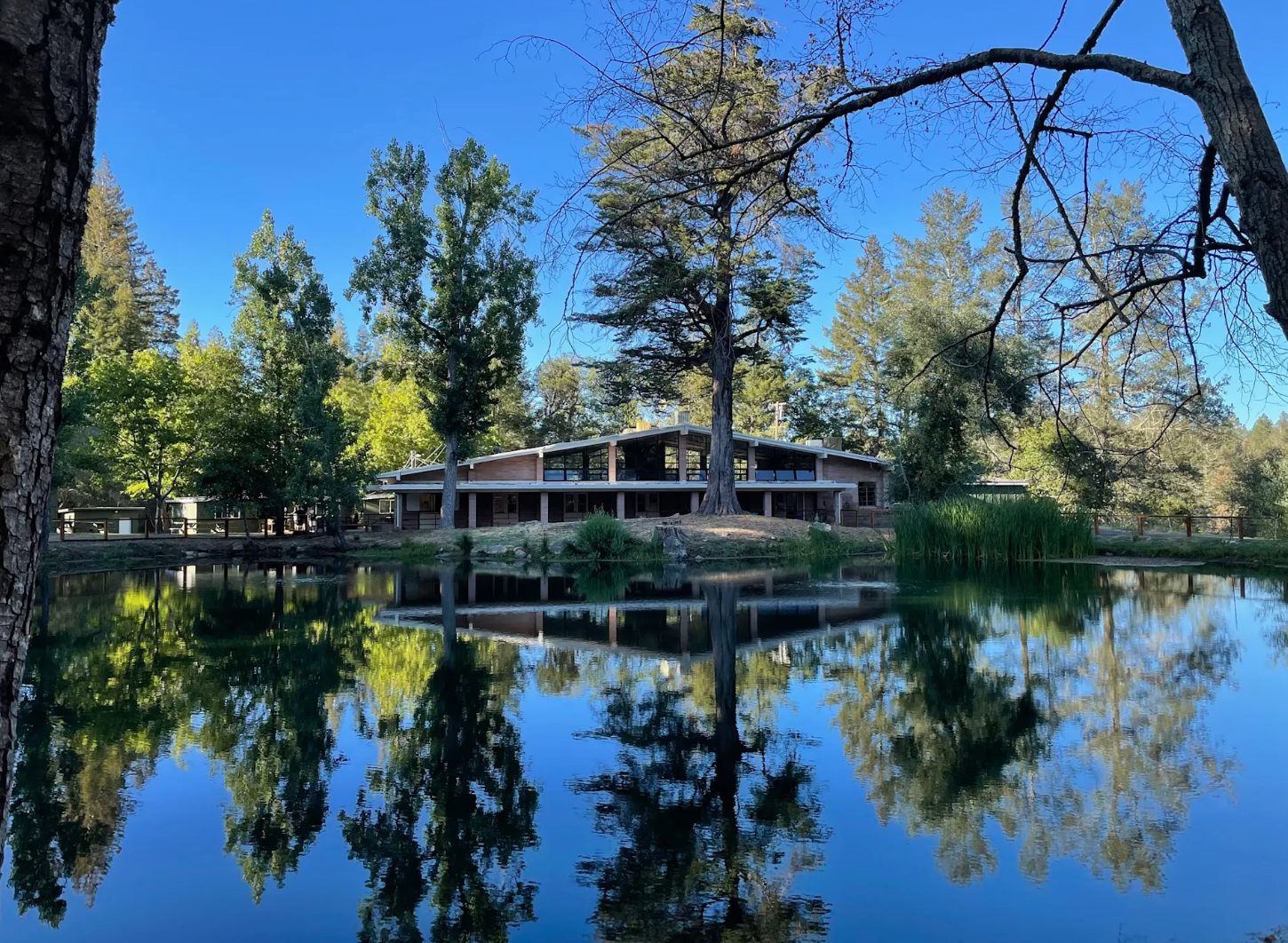 The Activities Hall at Enchanted Hills reflected in a calm lake with trees surrounding it